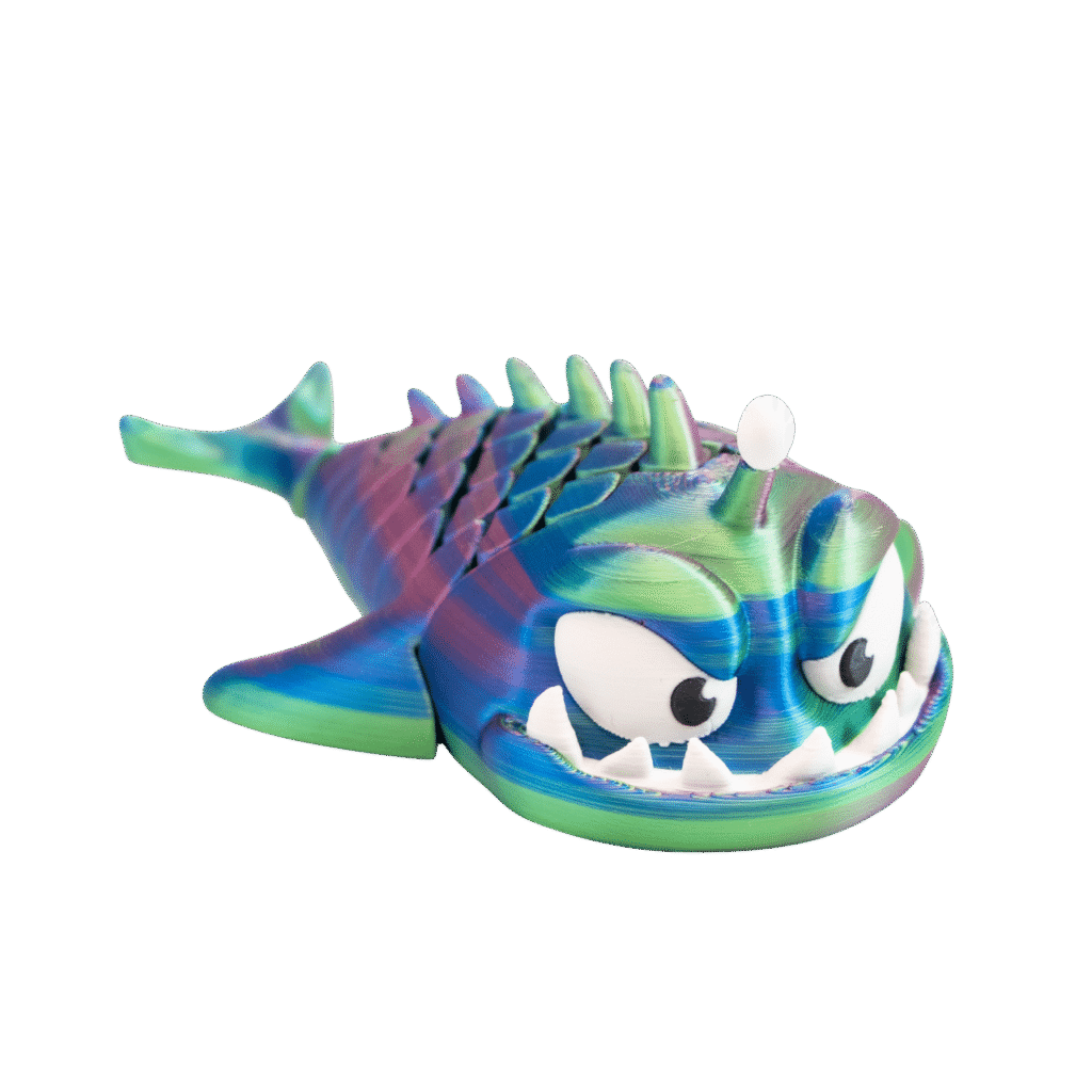 Rybeł – tęczowy brelok ryba z druku 3D leżący na boku. Morski potwór z szeroko otwartą paszczą pełną białych zębów, dużymi oczami i rzędem kolców na grzbiecie. Gradient kolorów: zielony, niebieski, fioletowy, różowy. Widoczne metalowe kółko do zawieszenia.