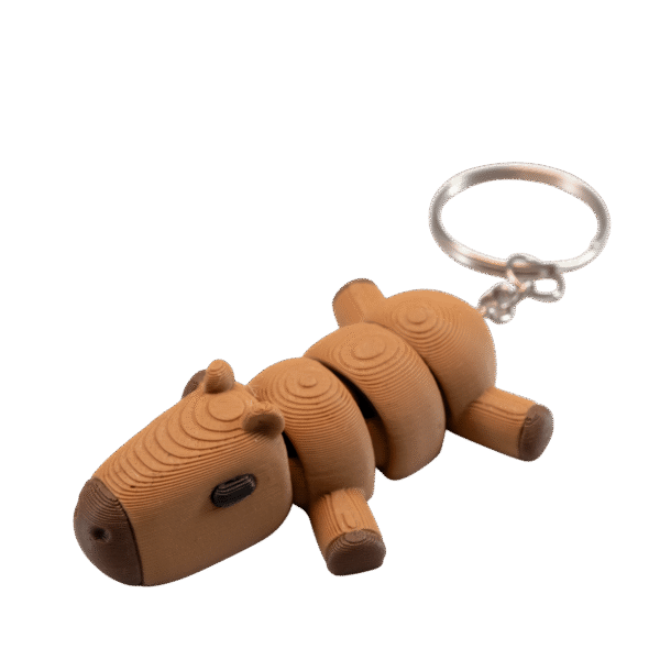 Kaprys – brelok kapibara z druku 3D leżący na boku. Beżowo-brązowa kapibara zbudowana z segmentów z widocznymi koncentrycznymi okręgami na powierzchni. Małe uszka, krótkie nóżki z ciemniejszymi stopkami, nos i oczy. Spokojna, relaksująca poza. Metalowe kółko do zawieszenia.