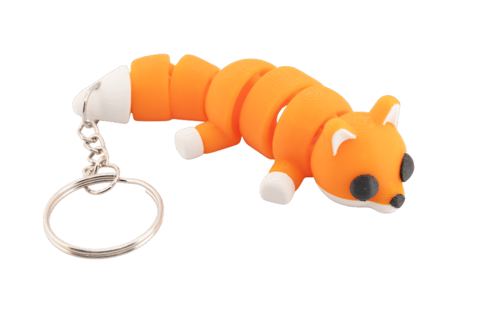 Miniz brelok lisek z druku 3D w intensywnym pomarańczowym kolorze, przedstawiony w pozycji leżącej z segmentowaną, giętką budową; lisek ma małe białe uszy, czarne okrągłe oczy, białą mordkę oraz białe łapki, a do tułowia przymocowany jest metalowy łańcuszek z kółkiem na klucze.