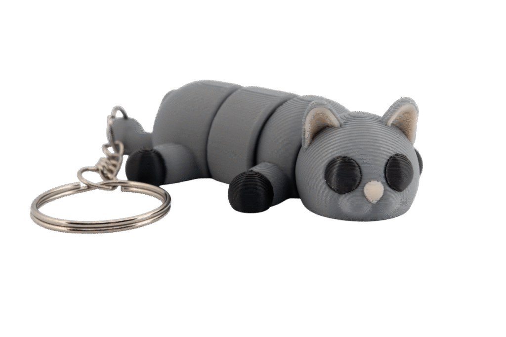 brelok kotek z druku 3D w kolorze popielatym, z segmentowym ciałem ułożonym w pozycji leżącej, z czarnymi oczami i czarnymi łapkami, jasnoróżowym noskiem, jasnoróżowymi wnętrzami uszu oraz metalowym kółkiem do kluczy po lewej stronie