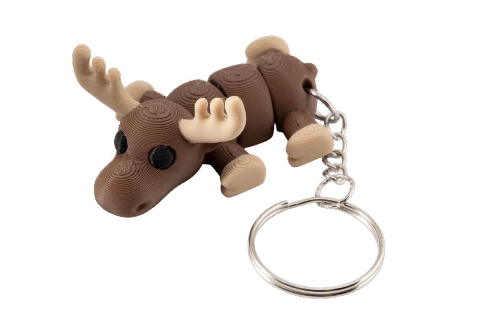 Brelok łoś z druku 3D w stylu miniz, w ciepłym brązowym kolorze, z dużymi kremowymi porożami i czarnymi oczami. Łoś leży w pozycji poziomej, z segmentowanym ciałem pozwalającym na ruch elementów. Do jego grzbietu przymocowany jest metalowy łańcuszek zakończony okrągłym, srebrnym kółkiem na klucze. Model ma gładką, warstwową fakturę charakterystyczną dla druku 3D.