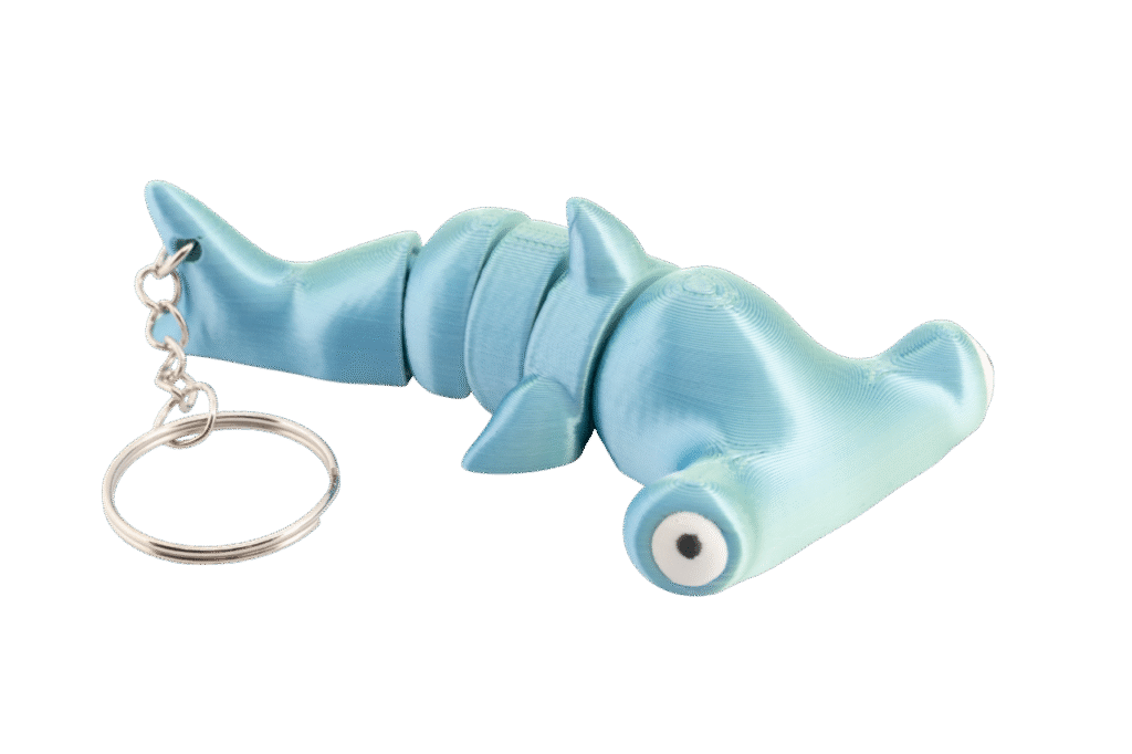 brelok rekin młot z druku 3d w kolorze jasnego turkusu, pokazany z boku w pozycji leżącej; ma segmentowane ciało z ruchomymi częściami, charakterystyczną szeroką głową młota, duże białe oczy z czarnymi źrenicami oraz metalowy łańcuszek i kółko do kluczy po lewej stronie