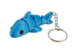 brelok rekin z druku 3d w intensywnie niebieskim kolorze, przedstawiający uroczego rekinka z dużą, gładką głową i okrągłym czarnym okiem, segmentowanym tułowiem z drobnymi, zaokrąglonymi płetwami po bokach oraz charakterystyczną płetwą ogonową; całość połączona z metalowym łańcuszkiem i kółkiem na klucze, leżąca na białym tle, w formie lekkiej, elastycznej zabawki-breloka
