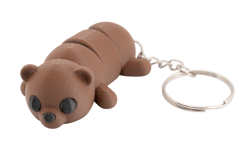 brelok miś z druku 3d w kolorze ciepłego brązu, przedstawiający leżącego misia o segmentowym, ruchomym korpusie; ma okrągłą głowę z małymi uszami, czarne oczy z delikatną teksturą warstw druku i mały czarny nosek, a z tyłu przymocowany jest metalowy łańcuszek z kółkiem do kluczy