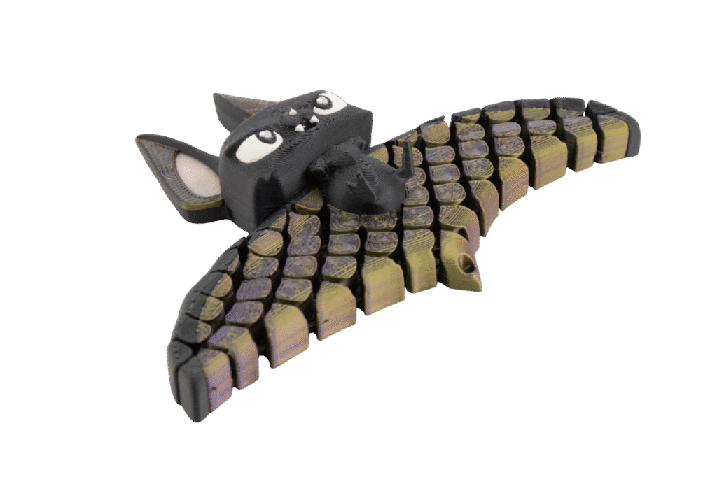 brelok nietoperz z druku 3d w kolorach czerni oraz przechodzącej fioletowo-złotej barwy na segmentowych skrzydłach, z dużymi białymi oczami i zwartą główką o wyrazistej, zabawnej mimice